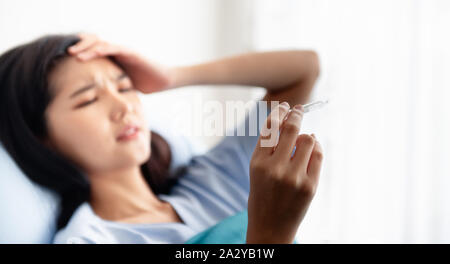 Eine junge Frau auf dem Bett des Krankenhauses verfangen war und mit Blick auf das Thermometer, sie krank, weil Fieber und Kopfschmerzen. Achoo, Grippe Saison. Stockfoto