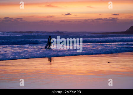 Der Surfer am Strand von Santa Cruz, der sein Surfbrett trägt, geht aus dem Wasser und macht eine Rock-on-Hand-Geste. Wolkiger Sonnenuntergang über dem Pazifik. Stockfoto