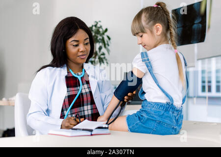 Jung und weiblich Afrikanischen Arzt einen Blutdruck ein kleines Mädchen. Kinderarzt Blutdruck messen von kleinen Mädchen patient Stockfoto