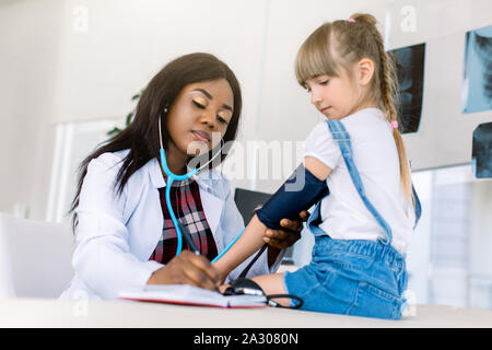 Wenig kaukasische Mädchen an Kinderarzt Büro Blutdruck messen. Junge afrikanische Frau Doktor Blutdruck messen Stockfoto