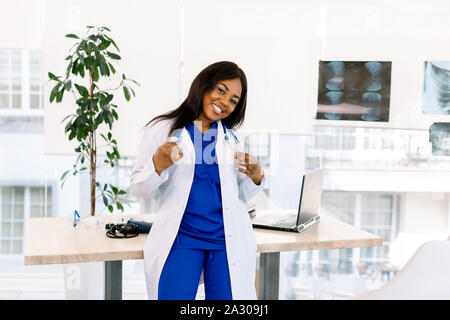 Nette junge afrikanische amerikanische Arzt Frau mit langem Haar mit medizinischen Einheitliche in glücklichen Ausdruck. Medizinische und Krankenschwester Konzept Stockfoto