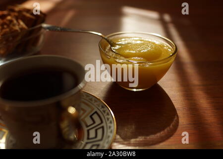 Honig in einem Glas Schale mit einem Löffel, eine Tasse Kaffee auf einer Untertasse, Cookies in einer Vase auf der sonnigen Tabelle. Stockfoto