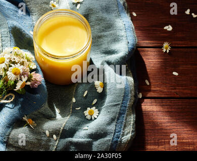 Geschmolzene Butter Ghee im lglass kann auf textered blau Textil mit Wiese Wildkräuter in der Nähe auf Holztisch, Heilung ayurvedische Heilmittel und sehr lecker Produk Stockfoto