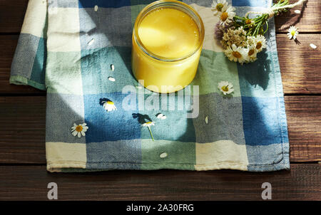 Geschmolzene Butter Ghee im Glas kann auf textered blau Textil mit Wiese Wildkräuter in der Nähe auf Holztisch, Heilung ayurvedische Heilmittel und sehr leckeres Produkt Stockfoto