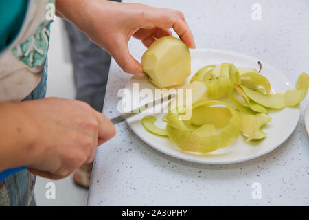 Weibliche Hände Peeling Haut aus der Green Apple mit einem Schälmesser mit Bambus platte Holztisch im Hintergrund. Direkt oberhalb der Schoß der Frau Peeling Stockfoto