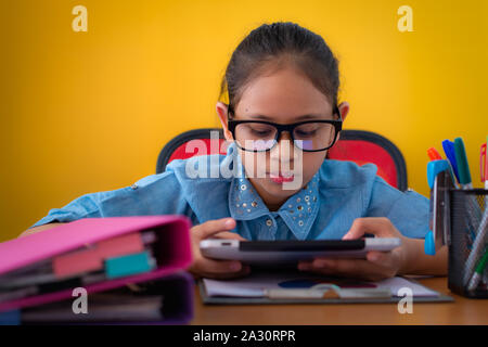 Süße Mädchen Tragen einer Brille mit Tablet-PC auf dem Schreibtisch isoliert gelben Hintergrund, Bildung Konzept. Stockfoto