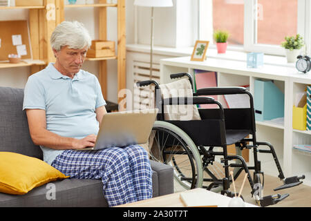Ältere Menschen in häuslicher Kleidung sitzt auf einem Sofa mit Laptop auf den Knien und Tippen auf er im Wohnzimmer ruht Stockfoto