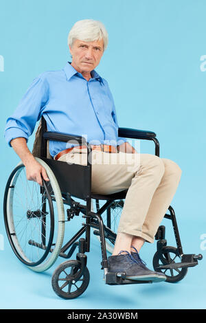 Portrait von behinderten älteren Mann sitzt im Rollstuhl und Kamera auf blauem Hintergrund isoliert Stockfoto