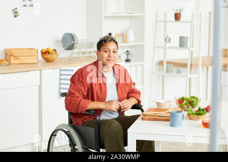 Portrait der Afrikanischen jungen behinderten Frau sitzt im Rollstuhl und mit Blick auf die Kamera in der Küche zu Hause. Stockfoto
