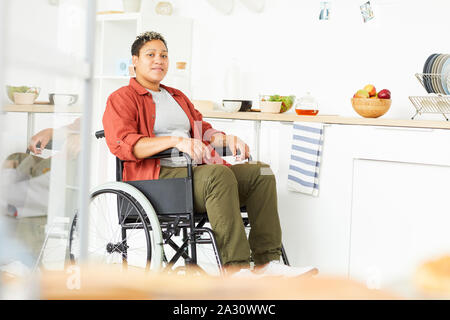 Porträt der Afrikanische junge Frau mit Behinderungen sitzt im Rollstuhl und mit Blick auf die Kamera in der Küche zu Hause. Stockfoto