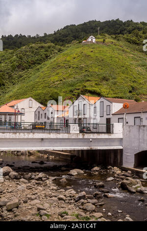 Fluss voller Steine, durch einen Wasserfall in den Bergen angelegt. Häuser eines Dorfes und einem Hügel mit einer kleinen Kapelle. Bewölkter Himmel. Da Terra Faial, Sao Mig Stockfoto