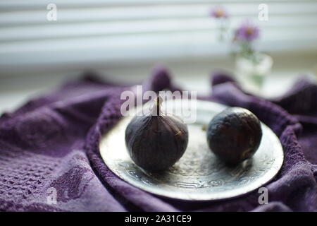 Zwei reife süsse Feigen auf einem Silbertablett. Tischdecke lila und violett Blumen auf der Fensterbank. Von der Seite. Stockfoto