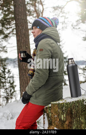 Süße Frau sitzt auf einem Baumstumpf im Wald, halten Sie eine Tasse mit einem heißen Getränk aus einer Thermoskanne und nachdenklich in die Ferne. Stockfoto