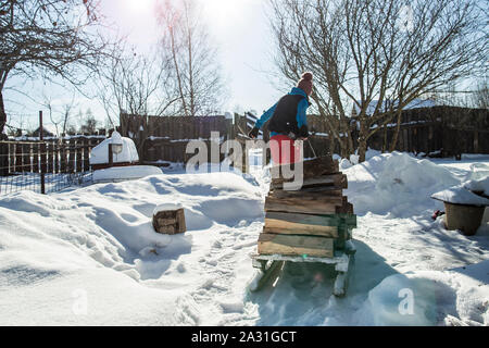 Eine junge Frau mit Schwierigkeit zieht eine alte hölzerne Schlitten mit Holz auf einem schneebedeckten Weg, in die Strahlen der Sonne, an einem frostigen Wintertag. Stockfoto