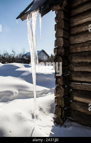 Eine leistungsstarke, lange transparente Eiszapfen hängen an den Rand des Daches des alten Hauses an und reicht bis zu den Schneeverwehungen. In der Hintergrundbeleuchtung von der Sonne, dem Stockfoto