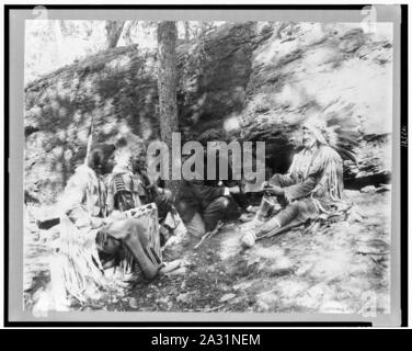 Ernest Thompson Seton, mit drei Schwarzfuß-Indianer, die zeigen, wie ein Feuer mit einem Bogen und einem Stock, Felsen und Bäume im Hintergrund starten Stockfoto
