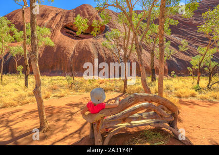 Touristische Frau mit breiten Hut auf Holzbank entlang Uluru Base Walk an der Südseite des Ayers Rock in den Uluru-Kata Tjuta National Park. Tourismus in