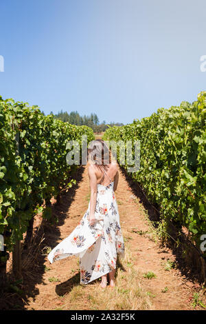 Schuss eine attraktive junge blonde Frau in einem weißen Sommerkleid tanzen und gerne Spinnen durch Reihen von Weintrauben. Stockfoto