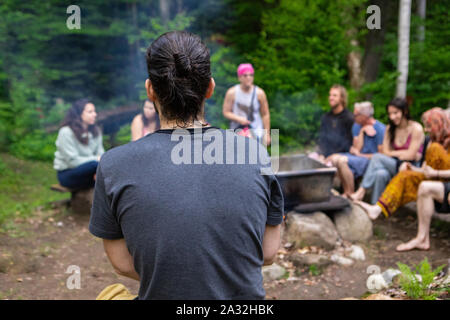 Eine Nahaufnahme und Rückseite des kaukasischen Kerl mit einem Haar bun- und grauen T-Shirt, sitzend um ein Campingplatz mit einer gemischten Gruppe von Menschen während einer mindfulness Retreat Stockfoto