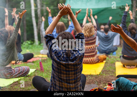 Eine generationsübergreifende Gruppe von Menschen tragen bunte Kleider sind von hinten gesehen, sitzen auf Yoga Matten in einer Waldlichtung, wie sie Meditation erleben. Stockfoto