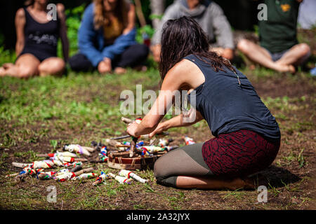 Einer böhmischen Stil Frau gesehen wird, Crafting heilige Objekte während einer woodland Retreat feiern Schamanismus und einheimischen Kulturen mit verschwommenen Menschen im Hintergrund. Stockfoto