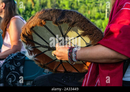 Eine Nahaufnahme auf den Arm und die Hand einer Person tragen ein rotes Hemd und bunten Perlen, die eine traditionell gestaltete native Drum mit pelzigen Kantenband. Stockfoto