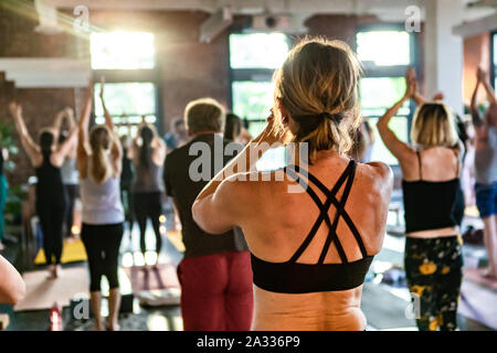 Ein kaukasischer Frau trägt einen schwarzen gegurtet Sports top Wird von hinten als Menschen der Praxis 108 Sonnengrüße in einem Fitnessstudio gesehen. Am Nachmittag scheint die Sonne durch die Fenster. Stockfoto