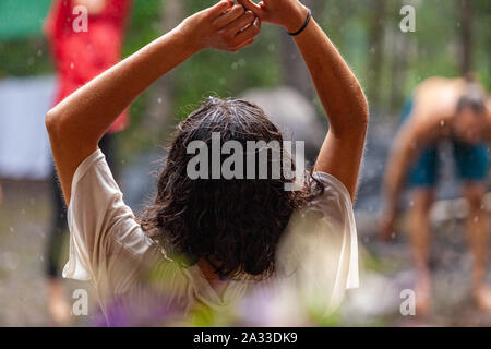 Eine junge Frau mit schwarzen Haaren von der Rückseite mit erhobenen Armen während einer schamanischen Ritual in ländlichen Woodland gesehen wird, unscharfe Teilnehmer sind im Hintergrund gesehen. Stockfoto