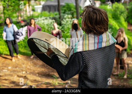 Eine 30 Jahre alte Schamane Kerl trägt einen bunten Schal ist von hinten gesehen, wie er eine mystische Trommel auf eine multiethnische Gruppe von Menschen während einer Rural Retreat spielt. Stockfoto