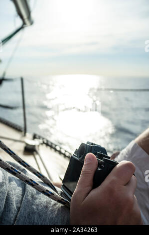 Ein Mann mit einer Kamera in der Hand entspannt sitzen auf dem Deck einer Yacht, auf einem unscharfen Hintergrund der Himmel, Meer und Sonne, ein warmer Sommertag. Stockfoto