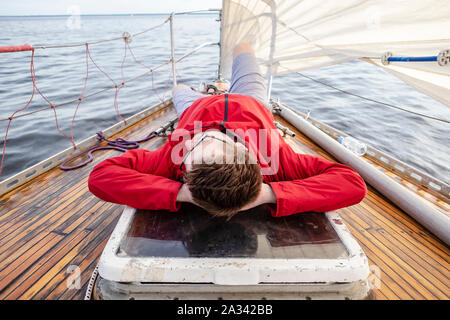 Ein Mann in einem roten Mantel und Gläsern entspannt auf dem Deck eines sich bewegenden Yacht, verschränkte die Arme unter seinem Kopf, vor dem Hintergrund des Meeres und des Himmels, o Stockfoto