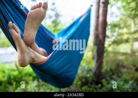 Die Füße des Menschen sind in der Nähe gesehen - aus einer Hängematte im Sommer Wald, auf einem unscharfen Hintergrund aus Pinien und einem See. Stockfoto