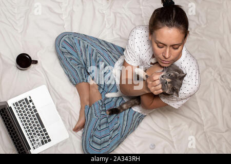 Eine Frau sitzt auf einer Decke und hält eine süße Katze in ihre Hände, neben ihr ist ein Laptop und eine Tasse Kaffee, in den frühen Morgenstunden. Stockfoto