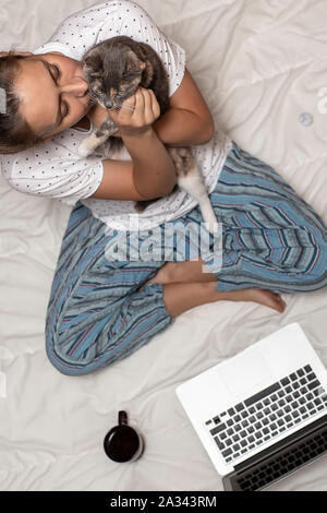 Eine Frau sitzt auf einer Decke und hält eine süße Katze in ihre Hände, neben ihr ist ein Laptop und eine Tasse Kaffee, in den frühen Morgenstunden. Stockfoto