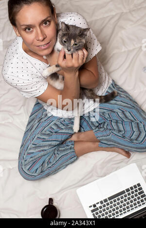 Eine Frau sitzt auf einer Decke und hält eine süße Katze in ihre Hände, neben ihr ist ein Laptop und eine Tasse Kaffee, in den frühen Morgenstunden. Stockfoto