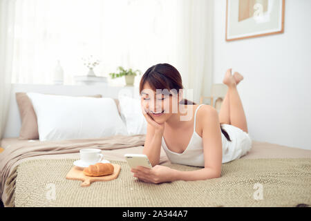 Fröhliche junge asiatische Frau mit Handy liegend auf Bett am Morgen Stockfoto