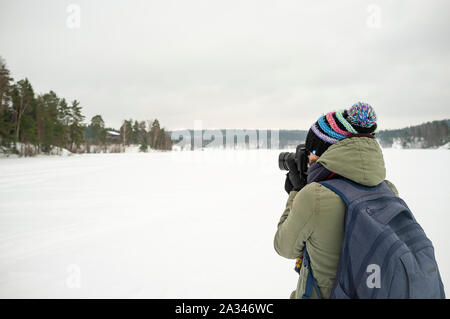 Eine Frau, die Fotografin in warme Kleidung hält eine Kamera in der Hand und nimmt Bilder der Winterlandschaft. Vor dem Hintergrund der See, Wald Stockfoto