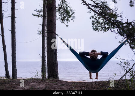 Der Mann entspannt in der Hängematte mit den Händen hinter seinem Kopf sitzen und genießen die Aussicht auf das Meer. Stockfoto