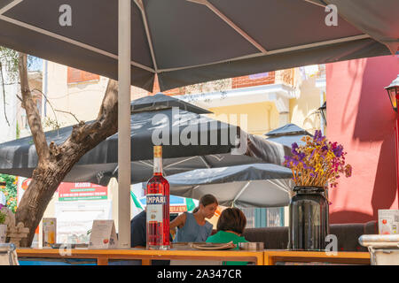 NAFPLIO GRIECHENLAND - 18 Juli 2019; griechische touristische Dorf von Nafplio street scene mit Campari Flasche auf Bar im Freien mit Sonnenschirmen und Menschen in backgroun Stockfoto