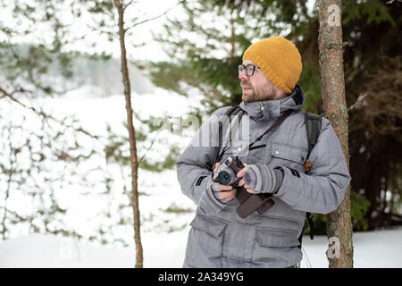 Ein bärtiger Mann mit Brille hält eine Kamera in den Händen und steht auf einem Baumstamm gelehnt eine junge Kiefer in einem Winter frostige Wald, gegen die hinterg Stockfoto