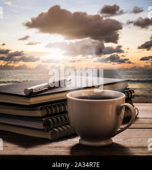 Heißer Kaffee mit Notebooks und Stift auf Holztisch mit Blick auf das Meer und den bewölkten Himmel in Abend Stockfoto