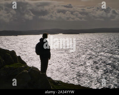 Ein einsamer, einzige Frau Walker, gegen die Spiegelung der Sonne auf dem Meer Silhouette - auf den Klippen von Muckle Roe in Shetland, Großbritannien Stockfoto