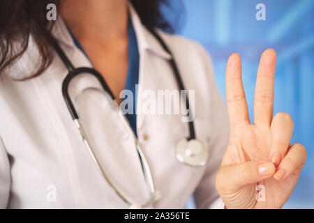 Junge schöne Arzt Frau mit Stethoskop über Krankenhaus Halle Hintergrund Blick in die Kamera zeigt die Finger tun Sieg unterzeichnen. Die Nummer zwei ist. Stockfoto