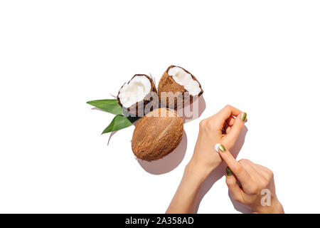 Blick von oben Frau Hände Auftragen kosmetischer Creme Neben natürlichen Kokosnüsse und frische grüne Blätter auf weißer Tisch, flach. Stockfoto