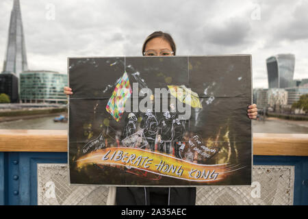 London, Großbritannien. Oktober 2019. Ein Aktivist aus Hongkong steht auf der Tower Bridge in London und hält ein großes illustriertes Poster mit Figuren und Regenschirmen mit Gasmasken und Slogans wie „Liberate Hong Kong“ und „Be Water“. Die Protestszene spiegelt die Solidarität mit der pro-demokratischen Bewegung in Hongkong von 2019 bis 2020 wider. Londoner Wahrzeichen wie das Shard- und Walkie Talkie-Gebäude sind im Hintergrund zu sehen. Penelope Barritt/Alamy Live News Stockfoto