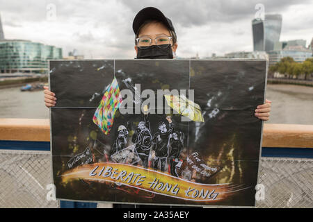 London, Großbritannien. Oktober 2019. Ein Aktivist aus Hongkong steht auf der Tower Bridge in London und hält ein großes illustriertes Poster mit Figuren und Regenschirmen mit Gasmasken und Slogans wie „Liberate Hong Kong“ und „Be Water“. Die Protestszene spiegelt die Solidarität mit der pro-demokratischen Bewegung in Hongkong von 2019 bis 2020 wider. Londoner Wahrzeichen wie das Shard- und Walkie Talkie-Gebäude sind im Hintergrund zu sehen. Penelope Barritt/Alamy Live News Stockfoto