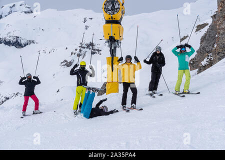 Junge Erwachsene im Winter, Schlange stehen am Hang das Tragen von Ski-, Snowboard-, Helm und Schutzbrille und Holding Sticks in die Luft mit Schnee berg im Hinterg