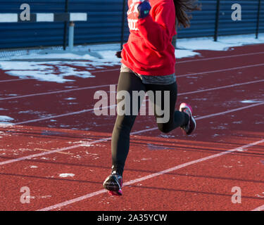 Ein High School Leichtathletik Läufer läuft auf einem vereisten und verschneiten Anschluss an einem sonnigen Nachmittag während der Praxis. Stockfoto