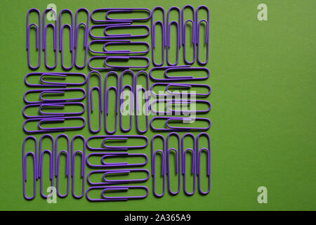 Lila Büroklammern in einem geometrischen Muster auf grünem Hintergrund mit einer isolierten roten und gelben Büroklammer zu einer Seite Stockfoto