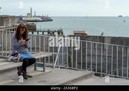 Eine Frau mittleren Alters auf einer Bank an einem View Point sitzen durch das Meer bei Ihrem Mobiltelefon oder Handy suchen Stockfoto